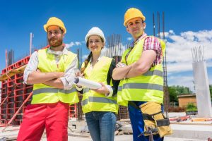 2 men wearing hard hats and safety vests at a job site and a woman wearing a hard hat and safety vest holding blueprints